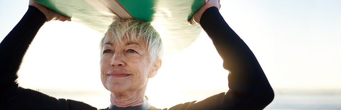 mature female surfer carrying surfboard over head.